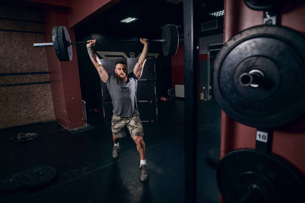 Muscular caucasian  bearded man holding barbell over his head doing barbell walking lunge exercise in crossfit gym. Weight plates in front. - Photo, Image