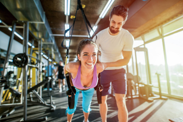 Determinate strong young personal trainer helping his female client to do TRX exercises correct. Client is looking happy and satisfied. - Photo, Image