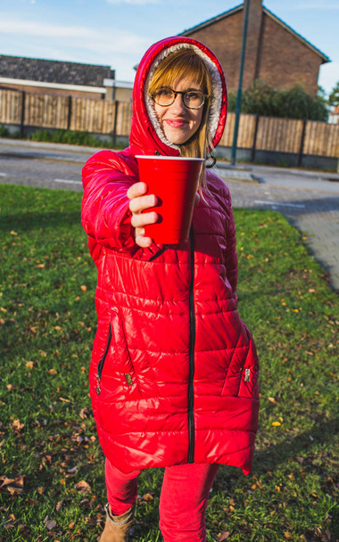 Χαμογελαστά ξανθιά κοπέλα με κόκκινα ρούχα, στέκεται πάνω σε ένα δρόμο και κρατώντας κόκκινη πλαστικό κύπελλο στα χέρια της. -Εικόνα - Φωτογραφία, εικόνα