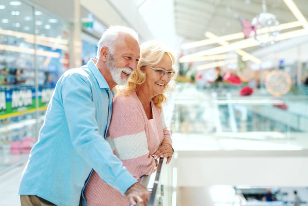 Smiling old Caucasian married couple leaning on the fence and looking down. Mall interior. - Foto, afbeelding