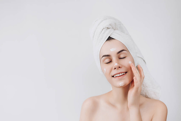 Skin care. A cute young  girl with a towel on her head puts a nourishing cream on her face. Causing moisturizer cream onto her face . Place for text - Valokuva, kuva