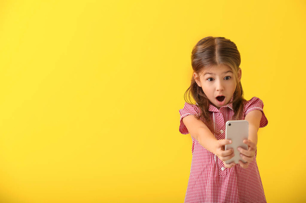Emotional little girl playing with smartphone on color background - Foto, Imagen