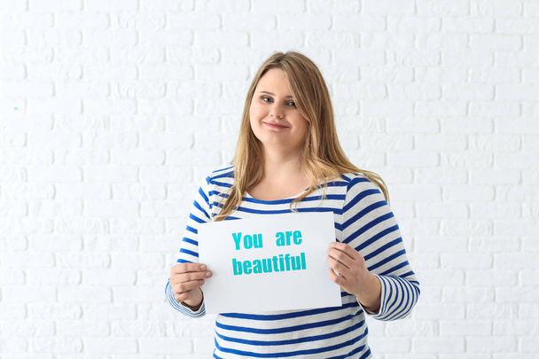 Beautiful plus size girl holding paper sheet with text YOU ARE BEAUTIFUL on white brick background. Concept of body positivity - Photo, Image