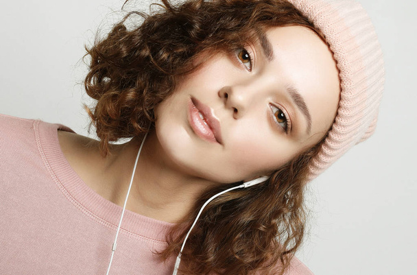 Portrait of a beautiful girl in headphones listens to music and smiles while standing on a white  background in a pink  sweatshirt and hat. Lifestyle concept. Close up. - Fotó, kép