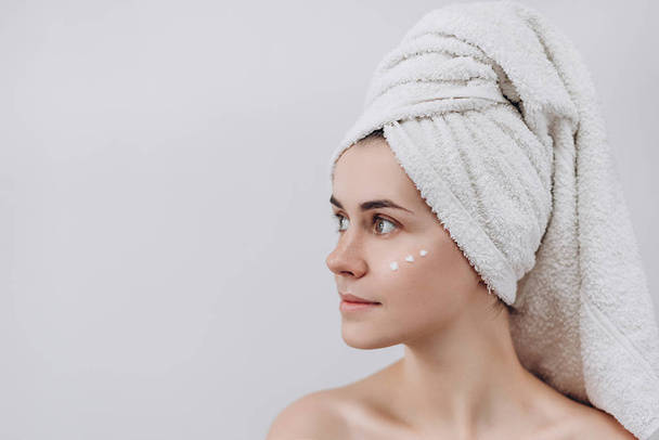 Woman with a towel on her head  looking above with face  moisturizer near eyes. Causing moisturizer cream onto her face. Intimate girl with perfect and healthy skin  on white background.  - Фото, изображение