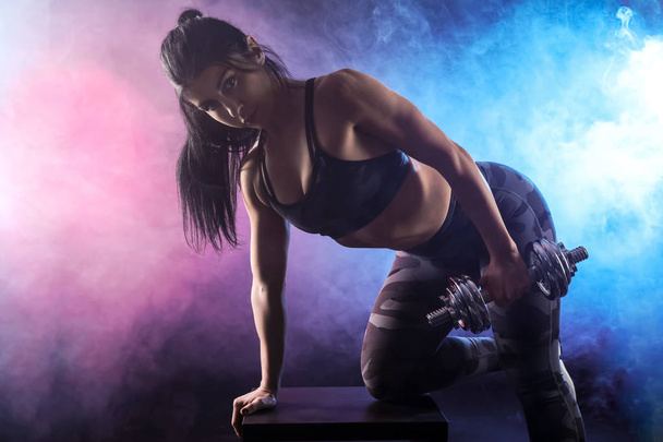 Sporty muscular woman with dumbbell in smoke on dark background - Fotoğraf, Görsel
