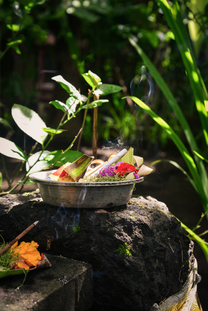 Traditional balinese offerings to gods in Bali with flowers and aromatic sticks on stone at garden - 写真・画像