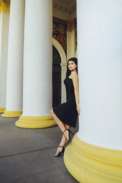 Young beautiful brunette girl in a black dress near the white columns - Foto, Imagem
