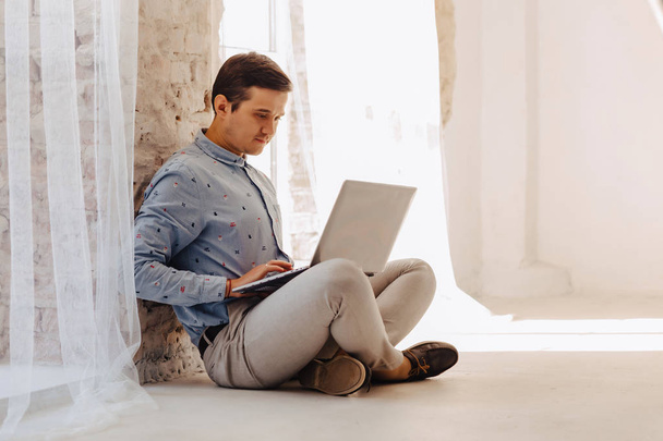 jonge stijlvolle man op de vloer werkt op laptop in lichte zonnige interieur, freelancer, notebook - Foto, afbeelding