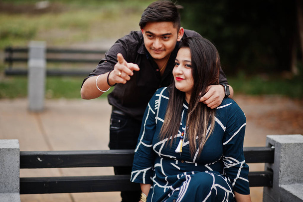 Love story of indian couple posed outdoor, sitting on bench together. Show finger - Photo, Image