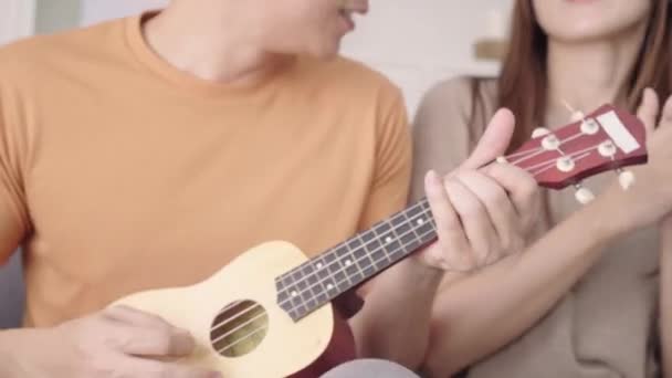 asiatisches Paar spielt Gitarre und singt gemeinsam im heimischen Wohnzimmer, süßes Paar genießt Liebesmomente, während es sich zu Hause auf dem Sofa entspannen kann. Lifestyle-Paar entspannt sich zu Hause. - Filmmaterial, Video