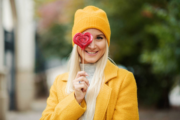 Saint Valentine's day concept. Fashion portrait blond young woman in yellow coat having fun with red lollipop heart over street background.  - Foto, Imagem