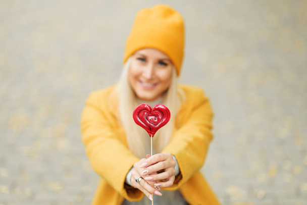 Saint Valentine's day concept. Fashion portrait blond young woman in yellow coat having fun with red lollipop heart over street background.  - Fotó, kép