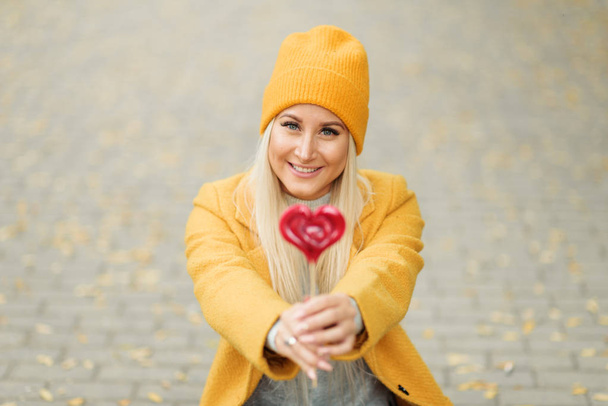Saint Valentine's day concept. Fashion portrait blond young woman in yellow coat having fun with red lollipop heart over street background.  - Zdjęcie, obraz