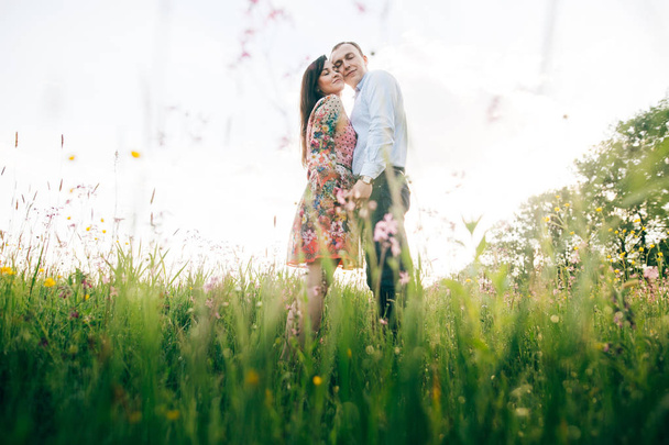 schönes junges Paar, das sich sanft in der Sonne auf einer frischen Frühlingswiese mit rosa Blüten umarmt. glückliche, stilvolle Familie, die sich im Sonnenlicht auf der grünen Wiese umarmt. Romantische Momente. Hallo Frühling - Foto, Bild