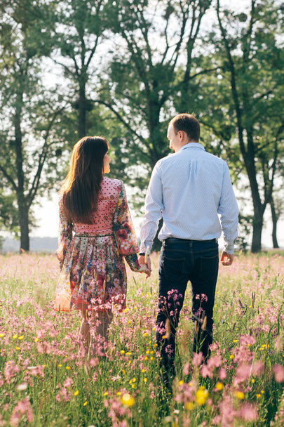 Gyönyörű fiatal pár kézen, és a gyaloglás a napsütés a tavaszi rét, rózsaszín virágokkal. Boldog család mosolyog a napfényben zöld mezőben. Romantikus pillanatok. Valentin nap - Fotó, kép