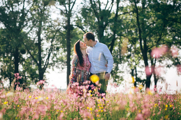schönes junges Paar Händchen haltend und im Sonnenschein auf der Frühlingswiese mit rosa Blüten spazierend. glückliche Familie lächelnd auf der grünen Wiese im Sonnenlicht. Romantische Momente. Valentinstag - Foto, Bild