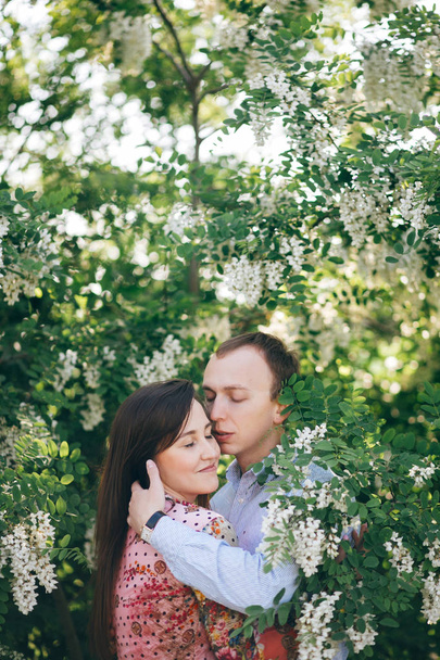 Beautiful young couple gently hugging in green leaves and white flowers in spring garden in sunshine. Happy family embracing at blooming acacia in sunlight. Romantic moments. - Photo, Image