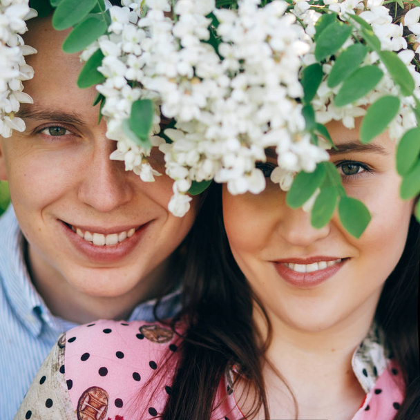 Poz ve bahar bahçe beyaz çiçekler altında gülümseyen güzel genç çift. Mutlu aile çiçek açan akasya ağacı güneş ışığı altında kucaklayan portreler. Romantik anlar. Sevgililer günü - Fotoğraf, Görsel