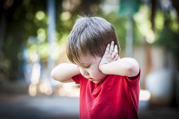 Un niño autista de 4 años con una camisa roja cerrando las orejas con las manos como protegiéndose del ruido. Concepto de autismo, síndrome de asperger, ruido fuerte, niño asustado, padres trauma de divorcio
 - Foto, Imagen