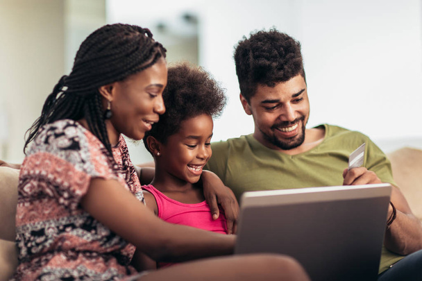 Cute little Afro-American girl and her beautiful young parents using a laptop and doing shopping online while sitting on a sofa at home. - Foto, Bild