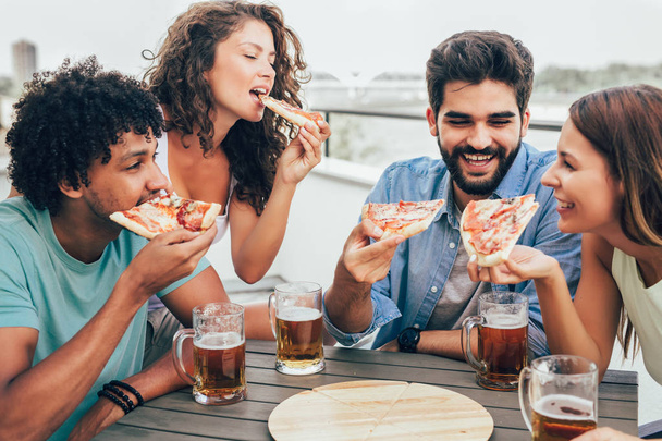 Ystävät nauttivat pizzasta. Ryhmä nuoria iloisia ihmisiä, jotka syövät pizzaa ja juovat olutta istuessaan papupusseissa rakennuksen katolla. - Valokuva, kuva
