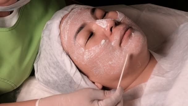 Az eljárás-és fiatalító tisztító a bőr kozmetika központjában. Spa kezelés egy nőt, kapott egy maszk szépségszalon. A kozmetikus, a kesztyű tisztítja a női arc eszközök. - Felvétel, videó