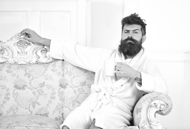 Man with beard and mustache enjoys morning while sitting on luxury sofa. Elite leisure concept. Man on sleepy face in bathrobe, drinks coffee, in luxury hotel in morning, white background - Valokuva, kuva