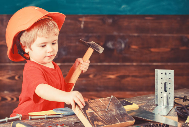 Toddler on busy face plays with hammer tool at home in workshop. Child in helmet cute playing as builder or repairer, repairing or handcrafting. Kid boy play as handyman. Handcrafting concept - 写真・画像