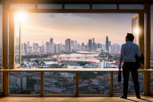 Concetto di visione, giovane uomo d'affari indossa comodo abito casual in piedi in sala di lavoro vicino alla finestra di vetro e guardando attraverso gli edifici moderni nello skyline
. - Foto, immagini