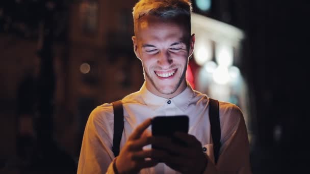 Young attractive man using smartphone standing on street of night town. Communication, travel, tourism, dating, business concept - Footage, Video