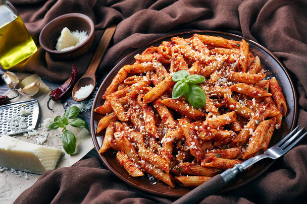 delicious penne arrabiata on a clay plate with fresh basil and grated pecorino cheese on a rustic table with brown cloth, fork and garetd cheese, view from above, close-up - Foto, Bild