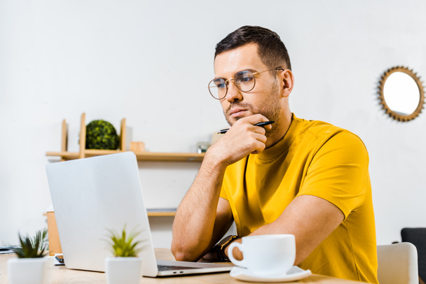 pensive man sitting in glasses and looking at laptop near cup of coffee - Foto, Imagem