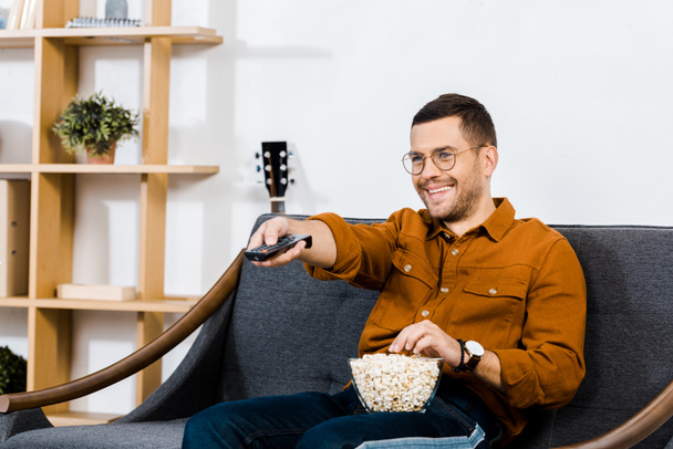 handsome man sitting on sofa with popcorn in bowl and holding remote control  - Φωτογραφία, εικόνα