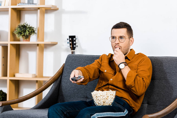 handsome man sitting on sofa, holding remote control and eating popcorn - Фото, изображение