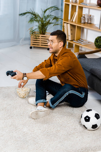 emocjonalną człowieka siedzi na dywanie w pobliżu popcornu podczas gry wideo  - Zdjęcie, obraz