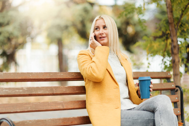 Young blond woman sitting in the city park and  drinking coffee to go, using mobile phone - 写真・画像