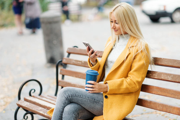 Young blond woman sitting in the city park and  drinking coffee to go, using mobile phone - Zdjęcie, obraz