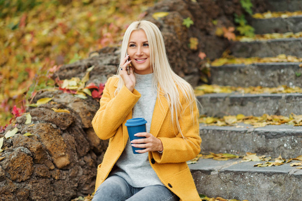 Young blond woman sitting in the city park and  drinking coffee to go, using mobile phone - Φωτογραφία, εικόνα
