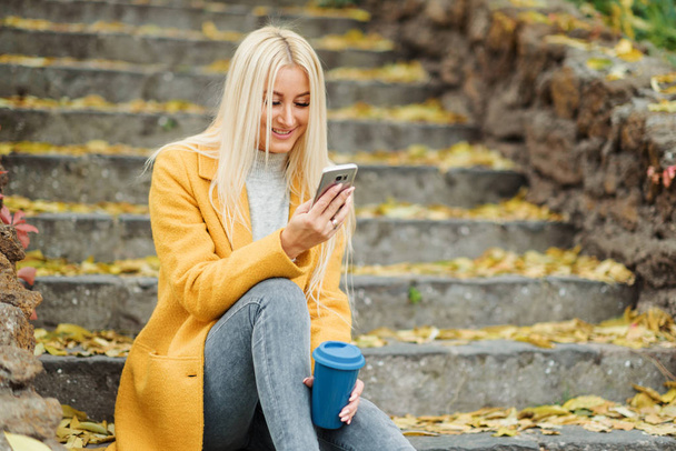 Young blond woman sitting in the city park and  drinking coffee to go, using mobile phone - Valokuva, kuva