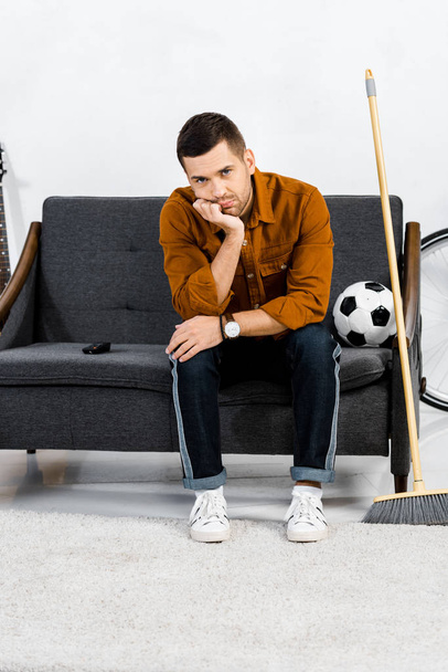 upset man sitting on sofa near broom and ball in modern living room  - Fotoğraf, Görsel