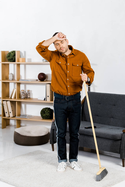 tired man standing with broom in modern living room  - Foto, Bild