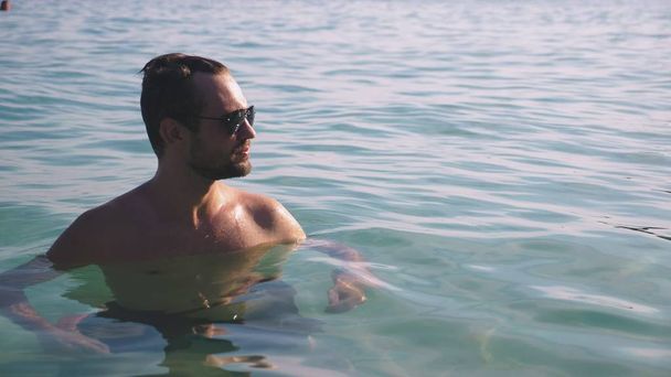Portrait of a handsome young man with a beard in sunglasses relaxed in crystal clear sea water - Φωτογραφία, εικόνα