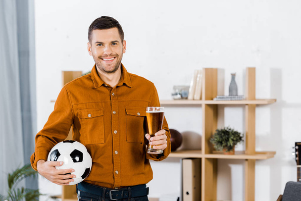 handsome man standing in modern living room while holding football ball and glass of beer - Фото, изображение