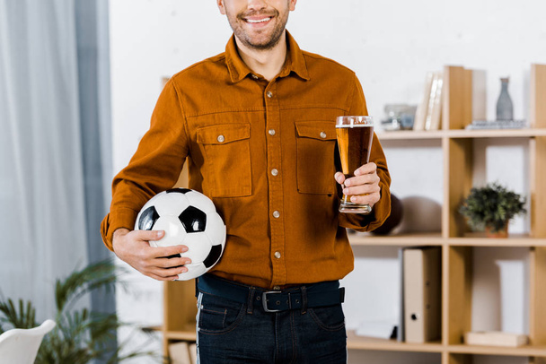 vista recortada del hombre de pie en la sala de estar moderna celebración de pelota de fútbol y vaso de cerveza
 - Foto, imagen