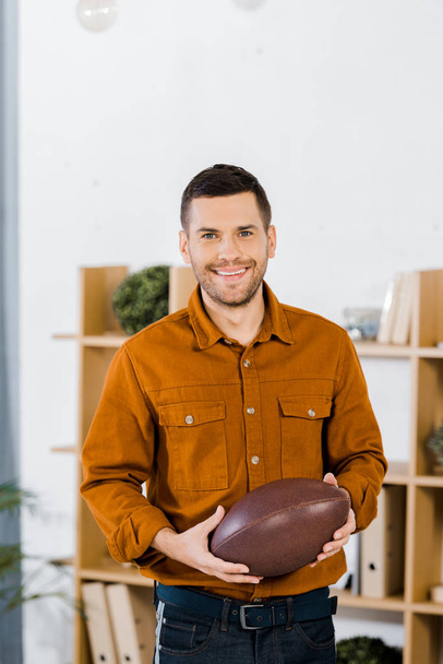 przystojny mężczyzna stojący w nowoczesnym salonie i trzymając piłkę futbol amerykański - Zdjęcie, obraz
