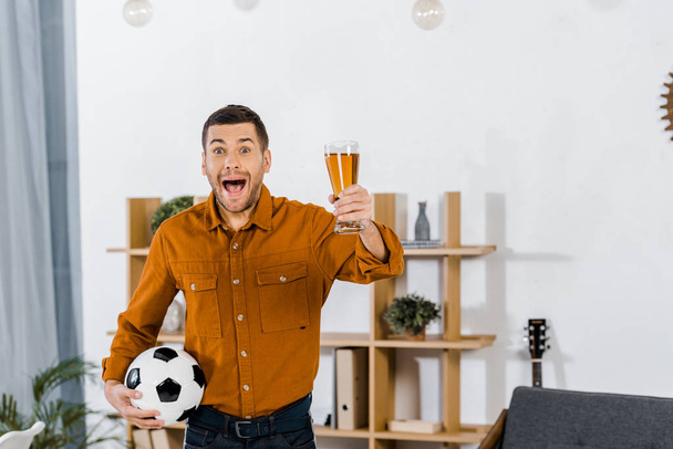 handsome man in modern living room screaming while holding football ball and glass of beer - Foto, Imagem