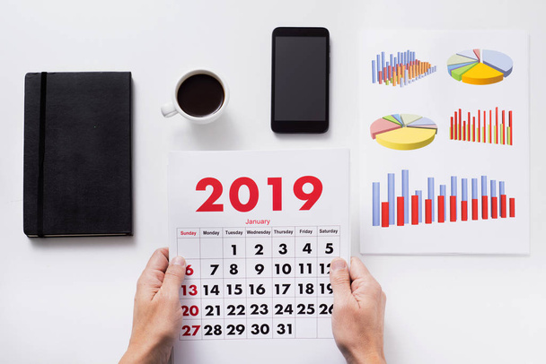 высокий угол обзора кавказца, сидящего за рабочим столом, соблюдающего календарь 2019 года, рядом с картами, чашкой кофе, смартфоном и блокнотом
 - Фото, изображение