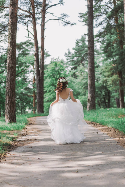 Brunette young bride with long beautiful curly hair. Woman with bridal elegant hairstyle decorated with fresh white flowers walking away from camera outdoors on summer sunny day. - Photo, Image