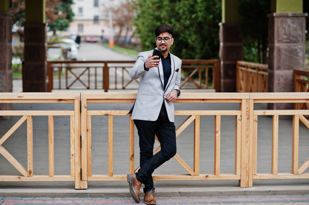 Stylish indian business man with bindi on forehead and glasses, wear on gray suit posed outdoor and looking at mobile phone. - Фото, изображение